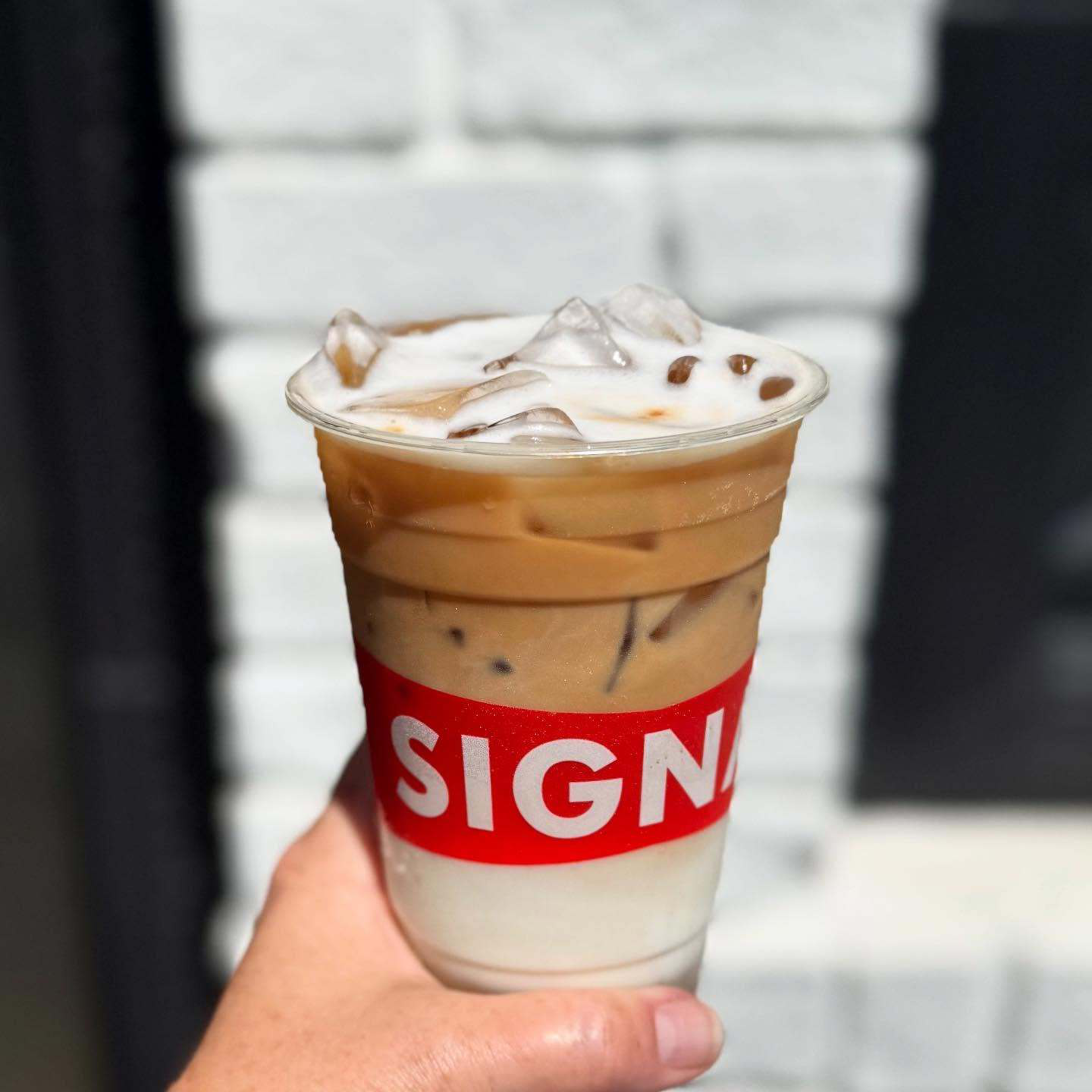 A hand holds a plastic cup of iced layered coffee with milk, topped with foam and ice cubes, featuring a red label partially reading “SIGN.”