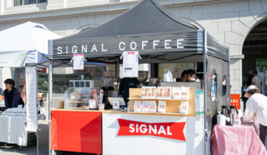 A black tent with "Signal Coffee" serves coffee and displays packaged coffee bags at a busy outdoor market stall.