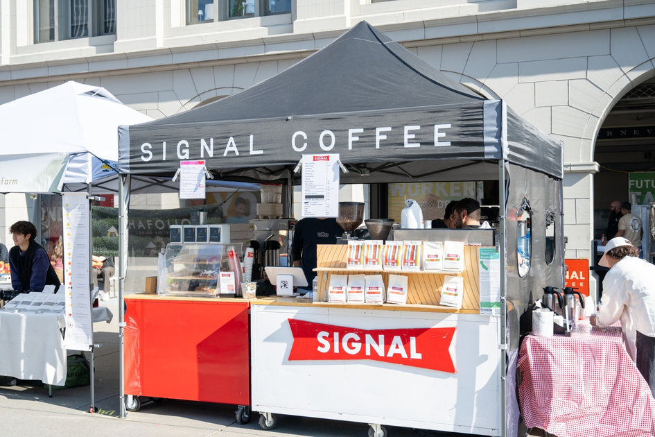 A black tent with "Signal Coffee" serves coffee and displays packaged coffee bags at a busy outdoor market stall.