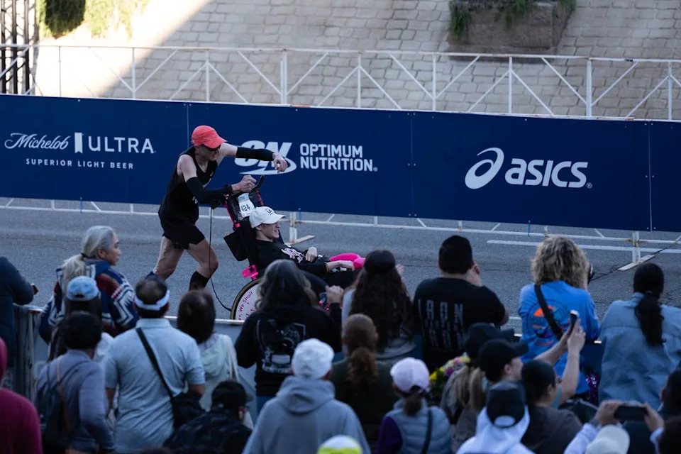 A runner pushes a participant in a racing chair past spectators near the finish line of the Los Angeles Marathon on Sunday, March 8, 2026, in Los Angeles.