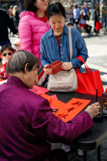 An elderly person writes Chinese calligraphy on red paper at a table while a woman stands nearby holding a red envelope. Other people are visible in the background.