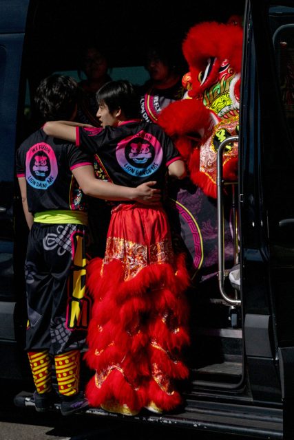 Two people in lion dance costumes stand in a van doorway, with one person wearing the red lion pants and others in matching team shirts.