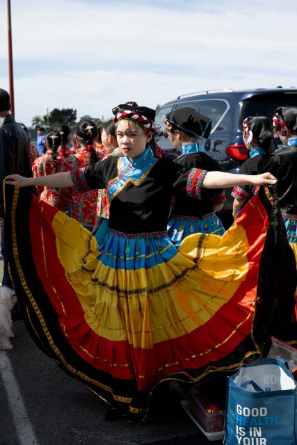 A young person in traditional colorful dress holds out their skirt at an outdoor event, surrounded by others in similar attire. A parked vehicle and a shopping bag are visible nearby.