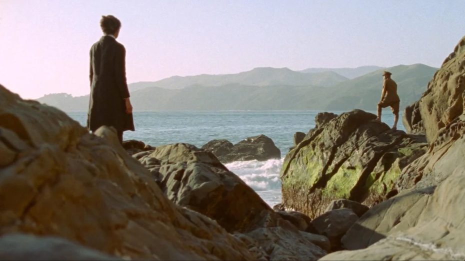 Two people stand on rocky cliffs by the ocean, facing the water, with mountains visible in the distance under a clear sky.