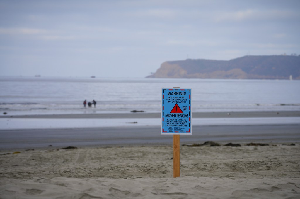 A sign on a beach warns, "BEACH WATER MAY CONTAIN SEWAGE AND MAY CAUSE ILLNESS."