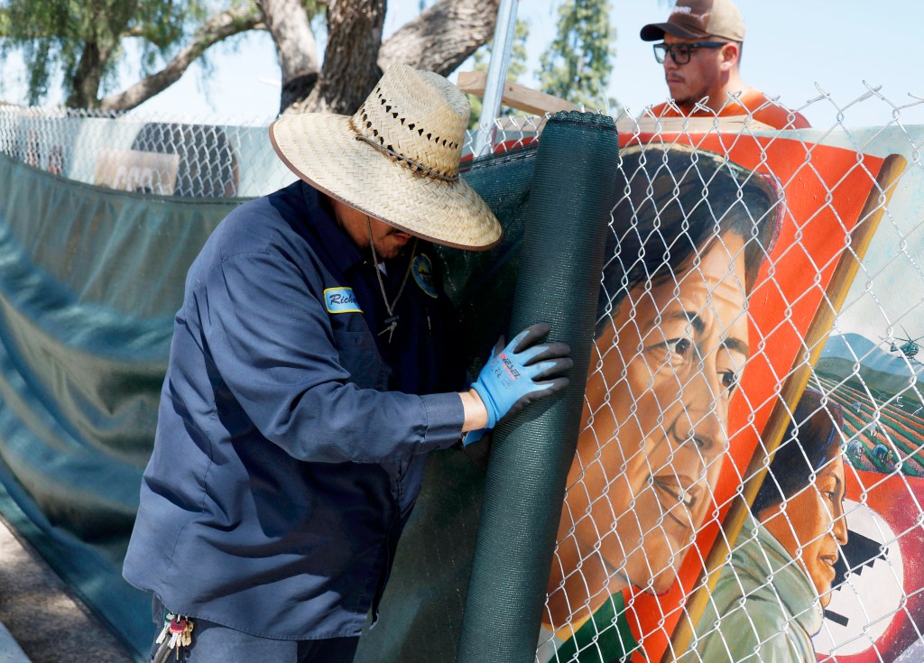 Two workers covering a mural of Cesar Chavez with a dark green screen.