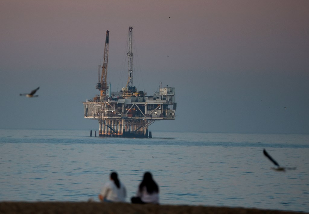 Offshore oil platform Esther in Seal Beach, California, with two people sitting on the beach in the foreground and gulls flying.
