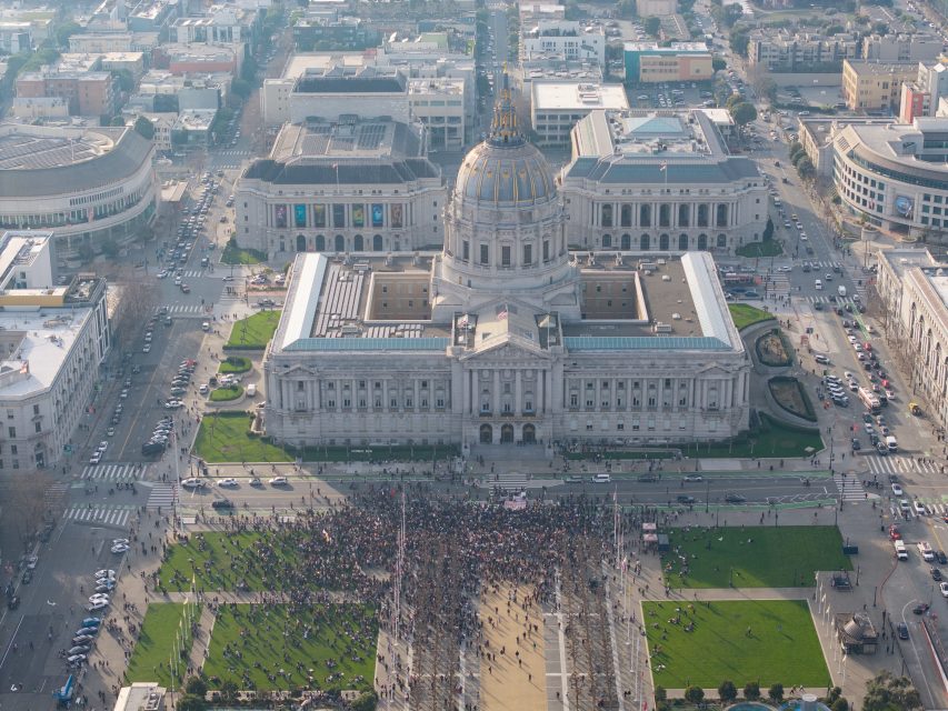 A large crowd gathers in front of a domed government building during an ICE walkout, with surrounding streets and cityscape visible from above.