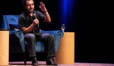 A speaker, Javier Zamora, gestures while seated on a blue chair, water glass nearby, under stage lights.