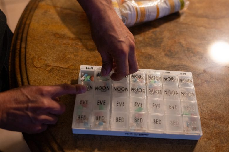 A person's hands interact with a weekly pill organizer on a table, focusing on the "NOON" compartment for Sunday.