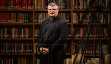 Paul Mariani stands in front of library shelves with chandelier in the foreground.