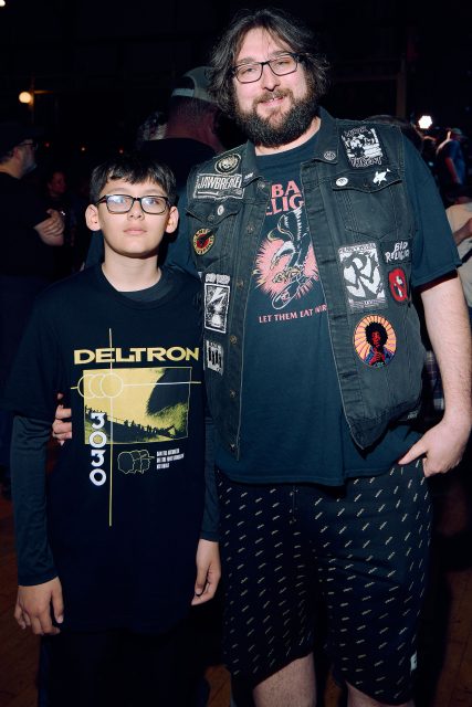 An adult and a child stand together at an indoor event; the adult wears a vest with patches, and the child wears a DELTRON t-shirt.
