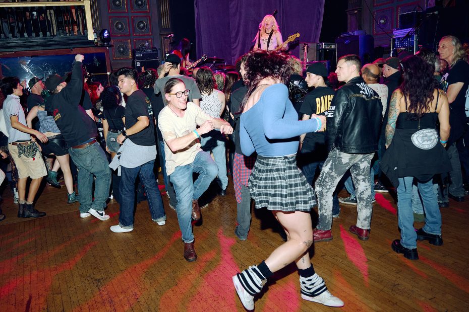 A crowd of people dance energetically in front of a stage where a band performs live music in a venue with wooden floors and decorative walls.
