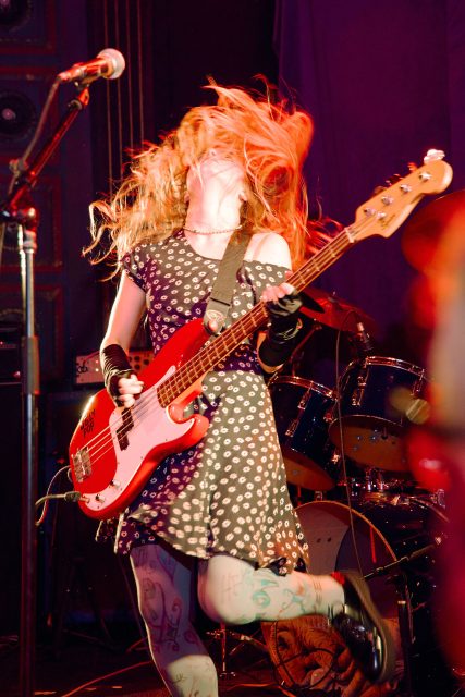 A musician with long hair plays an electric bass guitar energetically on stage, wearing a patterned dress and tights, with drums visible in the background.