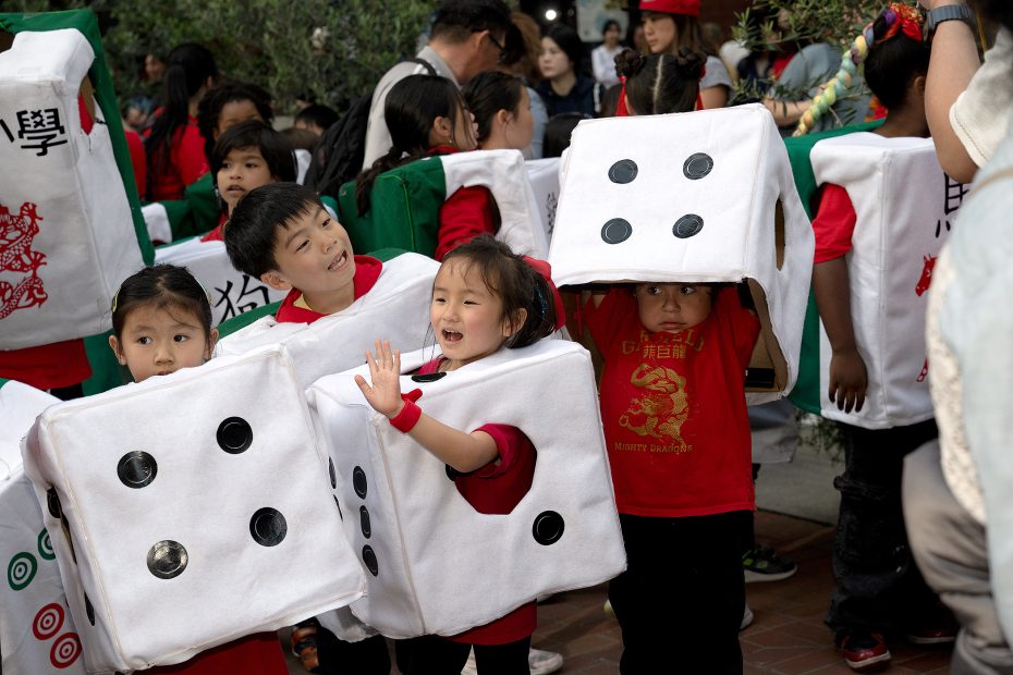 Several young children wearing large dice costumes and red shirts stand together in a crowd during an outdoor event.