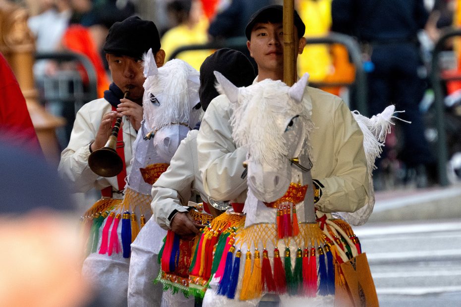 Two men wearing horse costumes and playing musical instruments participate in a parade, with colorful decorations and people blurred in the background.