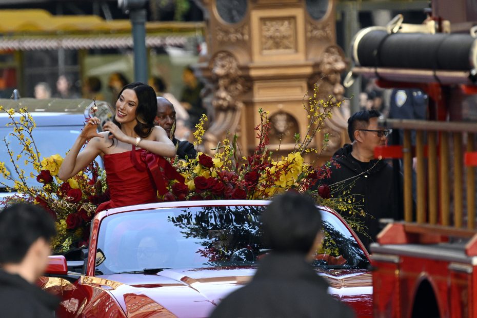 A woman in a red dress sits atop a red convertible decorated with flowers, holding a small object and smiling during a parade. People and vehicles are visible around her.