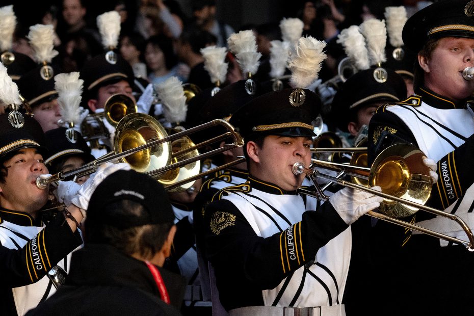 A marching band in black and white uniforms plays trombones in a crowded indoor setting, with a large audience in the background.