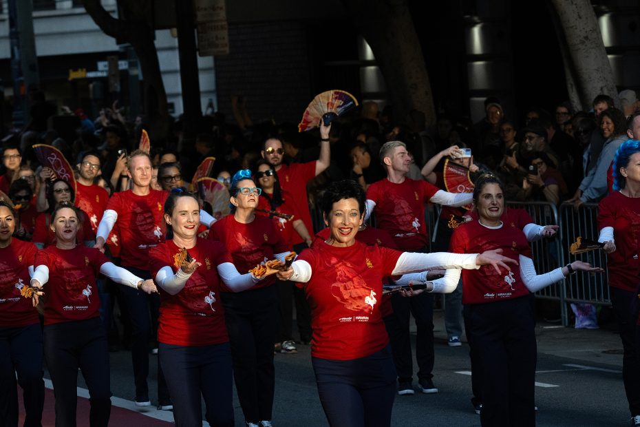 A group of people in matching red shirts and dark pants perform a coordinated dance in a street parade, with a crowd watching behind barricades.