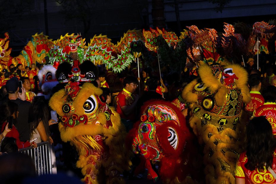 Performers in colorful dragon and lion costumes participate in a parade, surrounded by a crowd during a celebration at night.