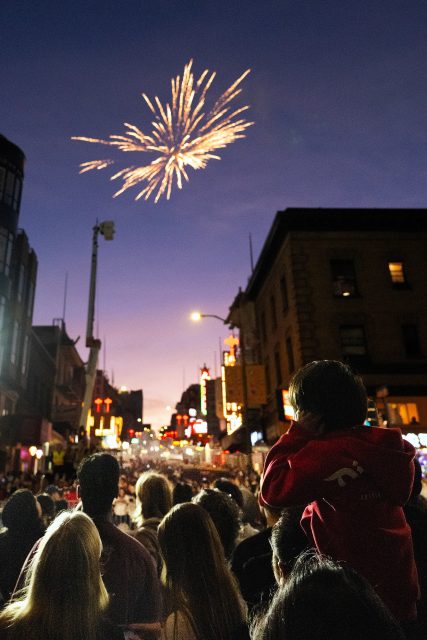 A crowd watches fireworks in the evening sky above a city street; a child sits on an adult’s shoulders in the foreground.