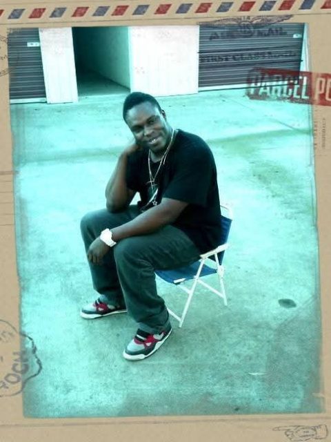 A man sits on a small folding chair outdoors in front of garage doors, smiling and resting his hand on his neck.