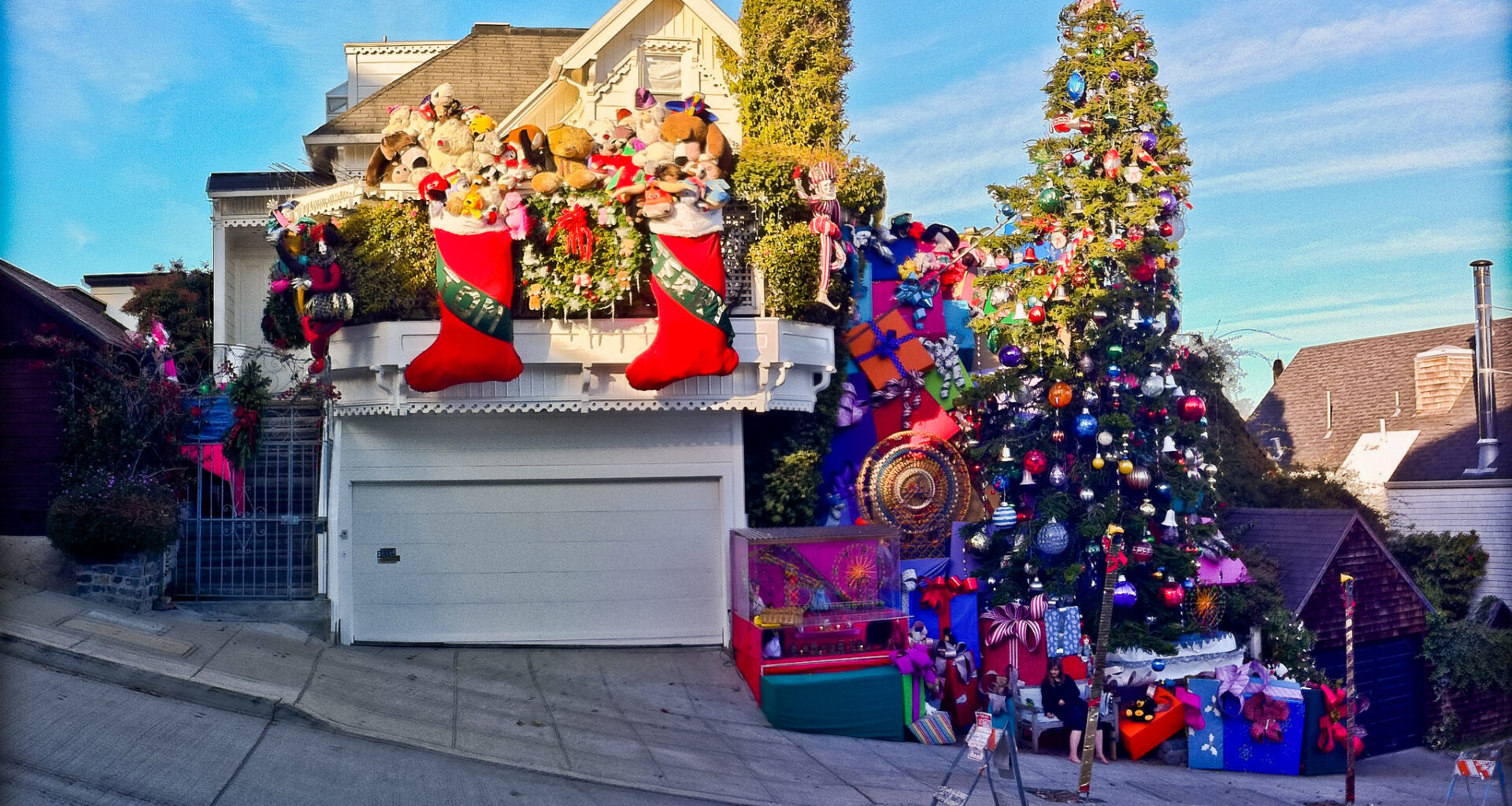 A house on a steep street is elaborately decorated for Christmas with large stockings, Santa figures, ornaments, a tall Christmas tree, and oversized gift boxes.