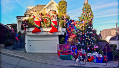 A house on a steep street is elaborately decorated for Christmas with large stockings, Santa figures, ornaments, a tall Christmas tree, and oversized gift boxes.