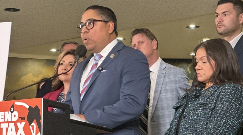A person in a blue suit speaks at a podium surrounded by colleagues. The group appears serious, likely during a press conference.