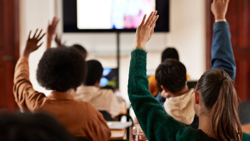 Rear view of multiracial students with hands raised in classroom at high school