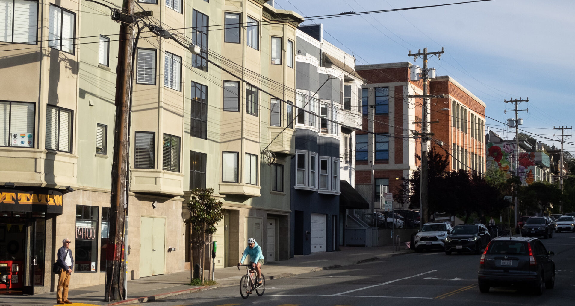 A person rides a bicycle through an intersection on a sunny city street lined with apartments, parked cars, and utility poles.