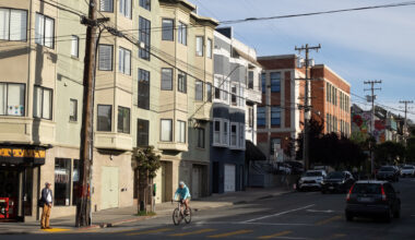 A person rides a bicycle through an intersection on a sunny city street lined with apartments, parked cars, and utility poles.