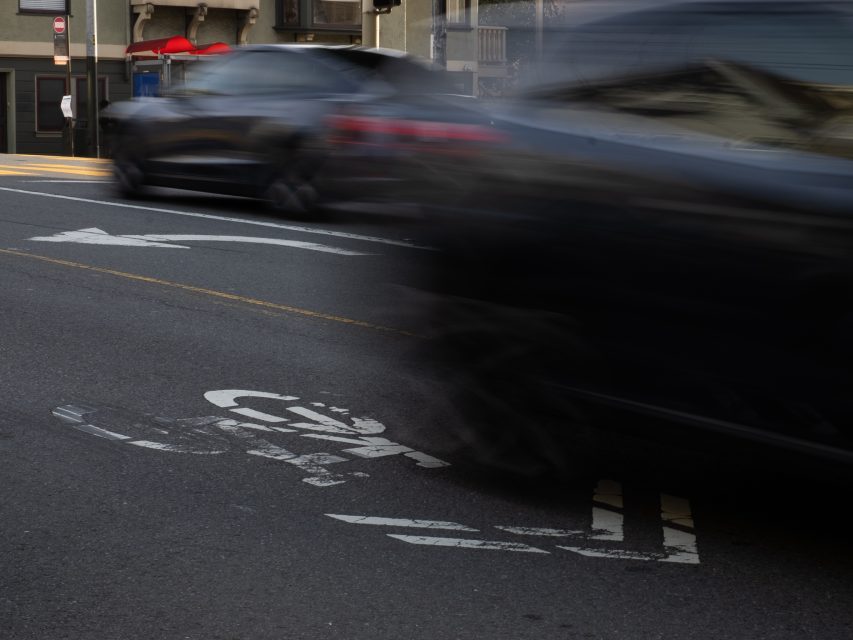 Two blurred cars drive past a faded bike lane marking on a city street, highlighting movement and urban traffic.