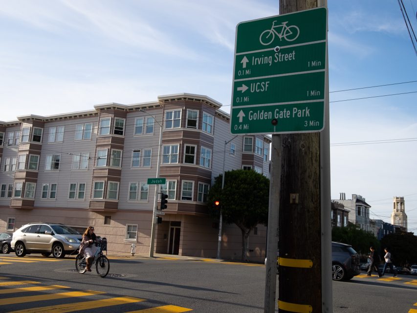 A street sign on a pole shows biking directions to Irving Street, UCSF, and Golden Gate Park. An apartment building and a cyclist crossing the street are in the background.