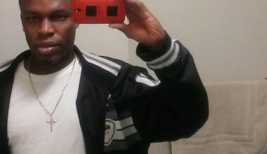 A man stands in front of a bathroom mirror taking a selfie with a red phone, wearing a black jacket, white shirt, and a cross necklace. A towel hangs on the wall behind him.