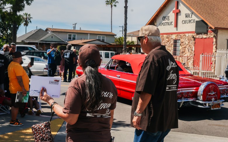 A pair wearing "Brown Image" car club shirts hold a commemorative lowrider stamp set on March 13, 2026 (Photo by Adrian Childress/ Special to Times of San Diego)