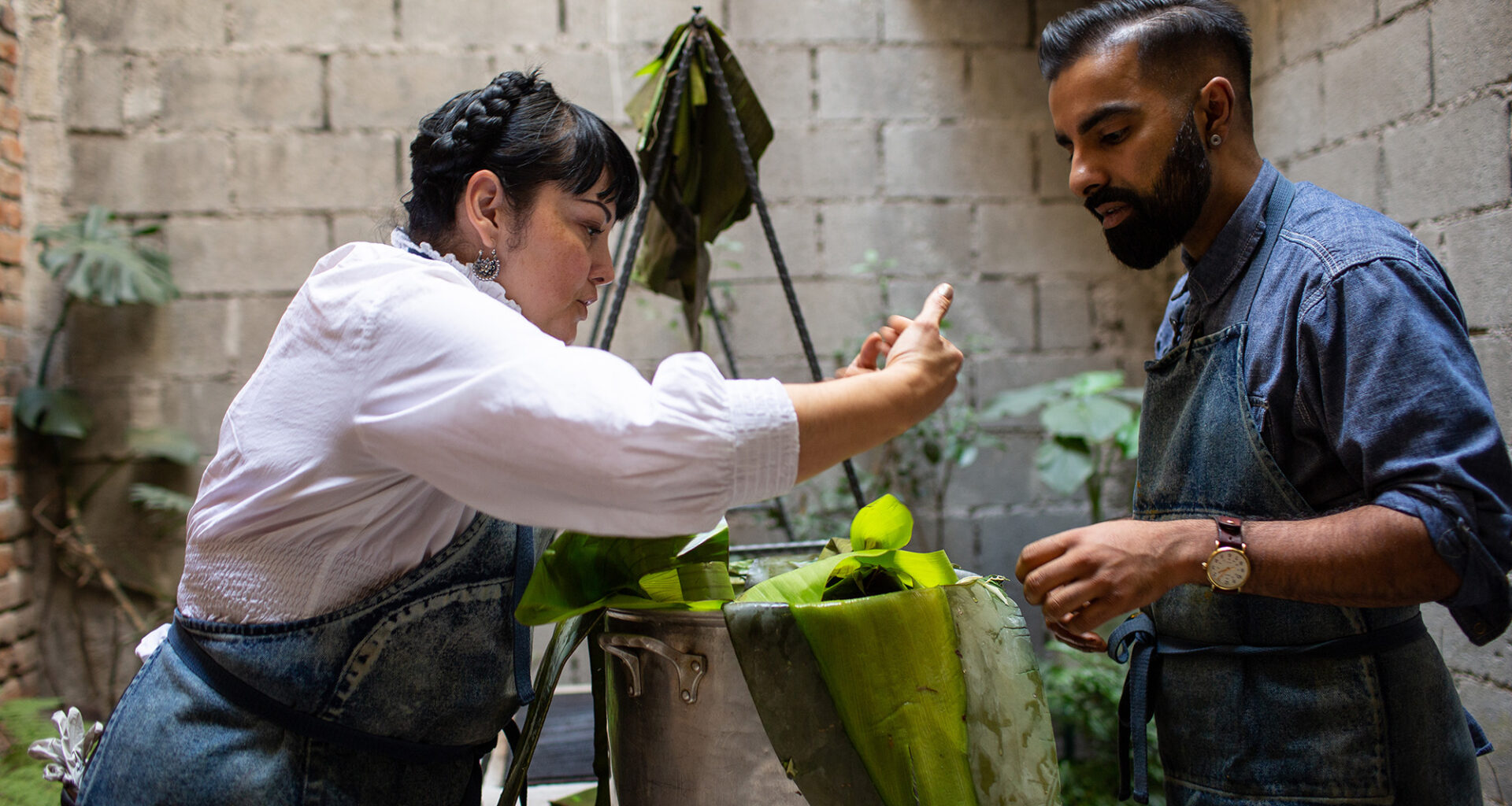 In an outdoor courtyard, a man and woman in blue aprons prepare banana-leaf tamales in a large pot.