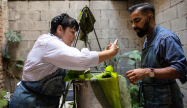 In an outdoor courtyard, a man and woman in blue aprons prepare banana-leaf tamales in a large pot.