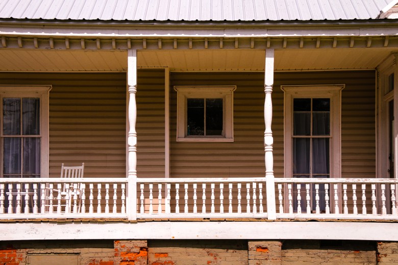 A wooden porch with white railings and columns features a single rocking chair in front of windows