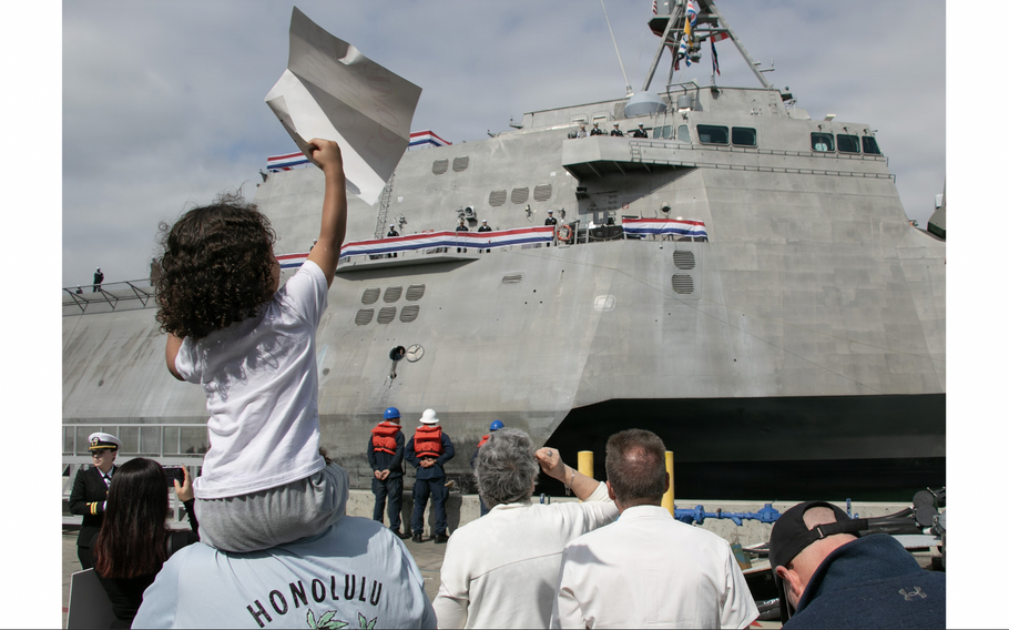 A child sits on the shoulders of an adult and waves a poster in the air while looking at a Navy ship.