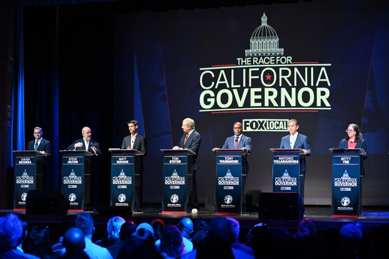 Seven candidates stand at podiums on stage during a California governor debate. A large screen behind displays "The Race for California Governor." Audience in view.