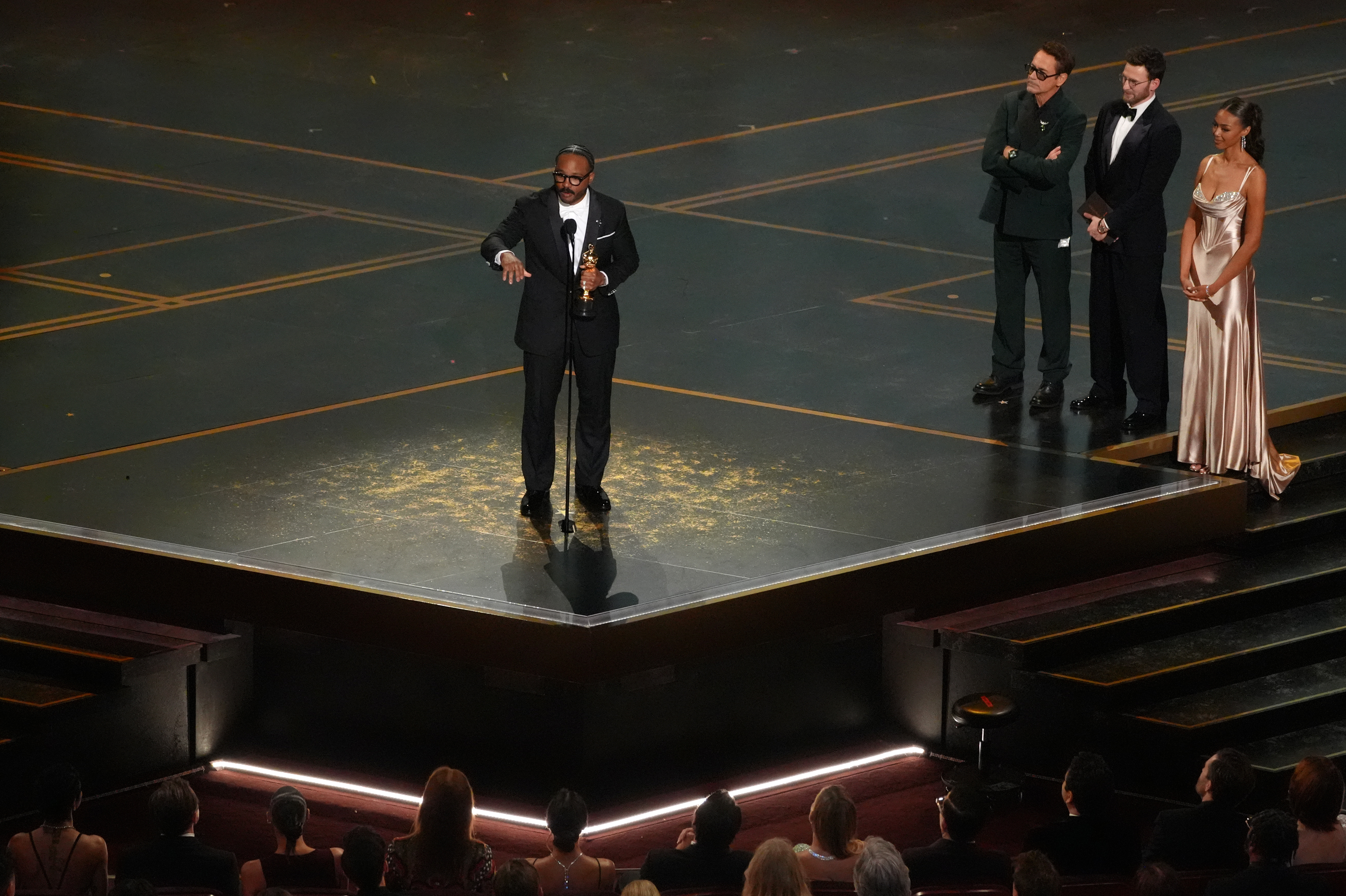 Ryan Coogler accepts the award for writing (original screenplay) for “Sinners” during the Oscars on Sunday, March 15, 2026, at the Dolby Theatre in Los Angeles. Robert Downey Jr., second right, and Chris Evans looks on from right.(AP Photo/Chris Pizzello)