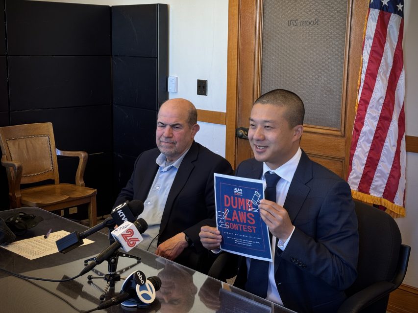 Two men sit at a table with microphones, one holding a “Dumb Laws Contest” poster; an American flag is visible in the background.