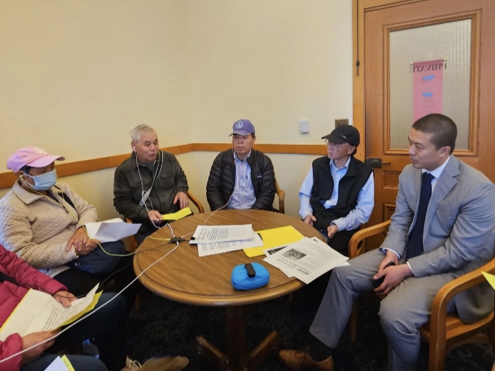 Six people sit around a small round table in a meeting room, discussing documents and papers, with one person wearing a mask.