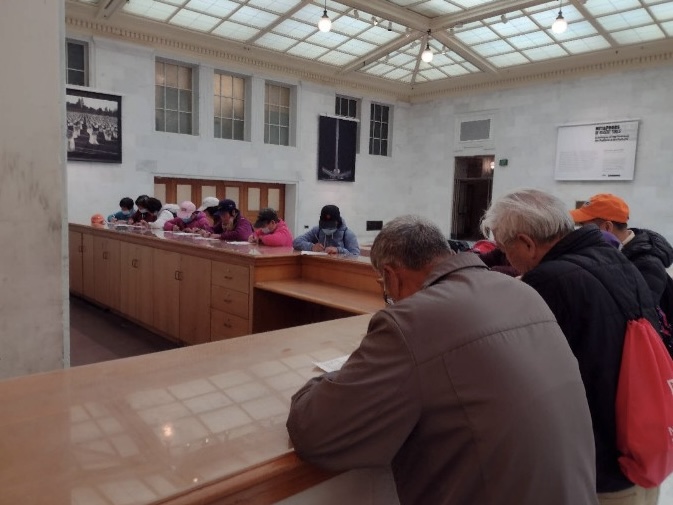 People are filling out forms or paperwork at a long wooden counter in a spacious, well-lit room with high ceilings and large windows.