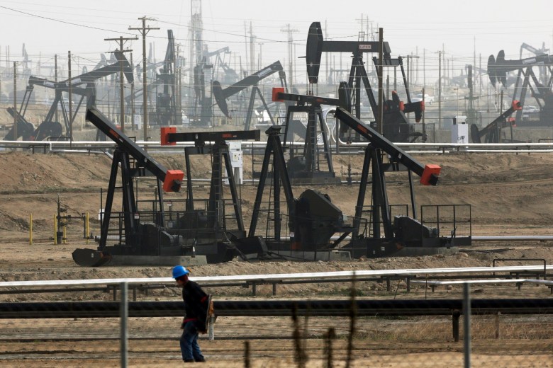 Oil pumps dot a dusty landscape, silhouetted against a hazy sky. A person in a blue hard hat walks in the foreground.