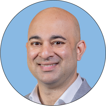 A bald man wearing a light gray suit jacket and white shirt, smiling, in front of a blue background.
