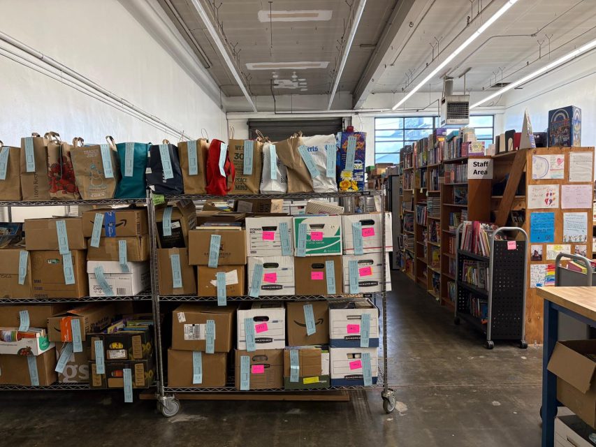 Shelves filled with cardboard boxes, bags, and books are neatly organized in a well-lit room with concrete floors and fluorescent lights. A staff area and more bookshelves are visible in the background.