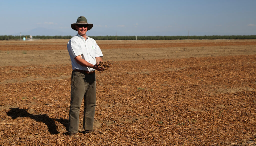 Brent Holtz, wearing dark heans, a short sleeved blue shirt and a cowboy hat, stands in an empty farm field that is strewn with the chips of old almond trees