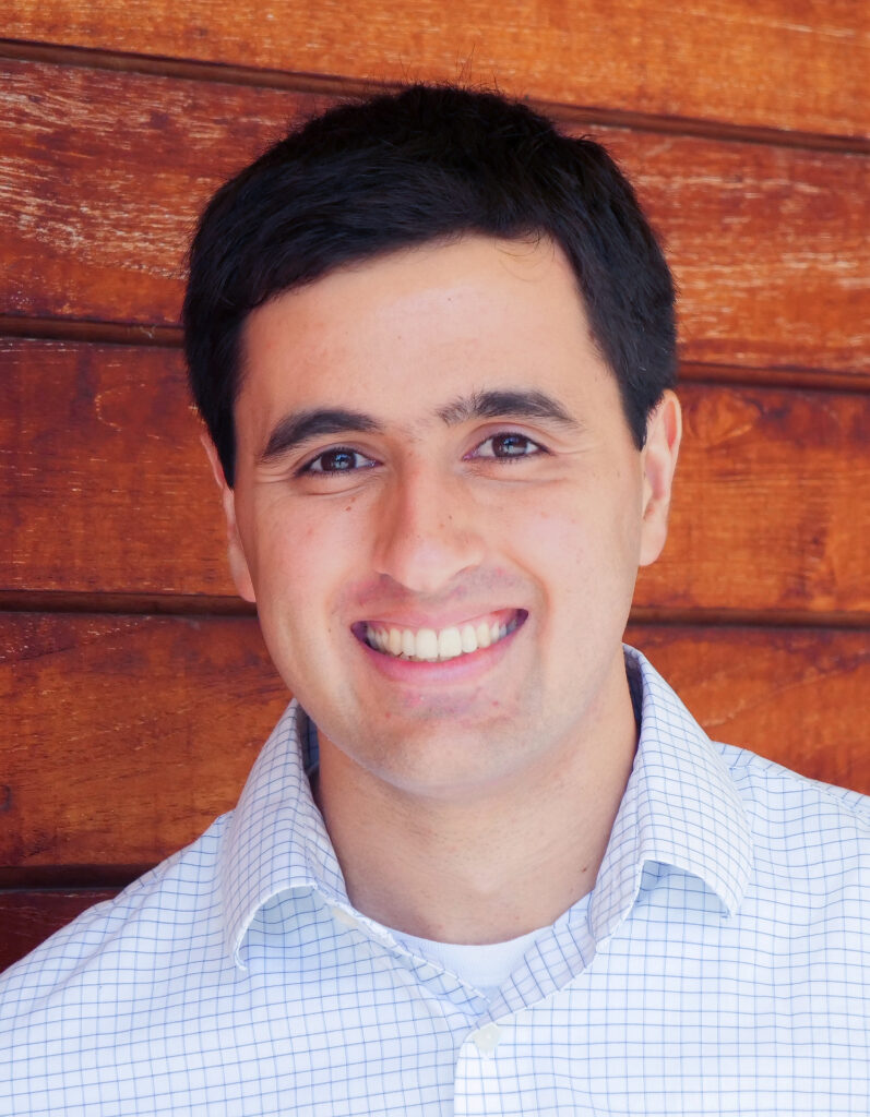informal vertical headshot of Brett Fischer, in an open-collar shirt, standing against a wall of wood planking
